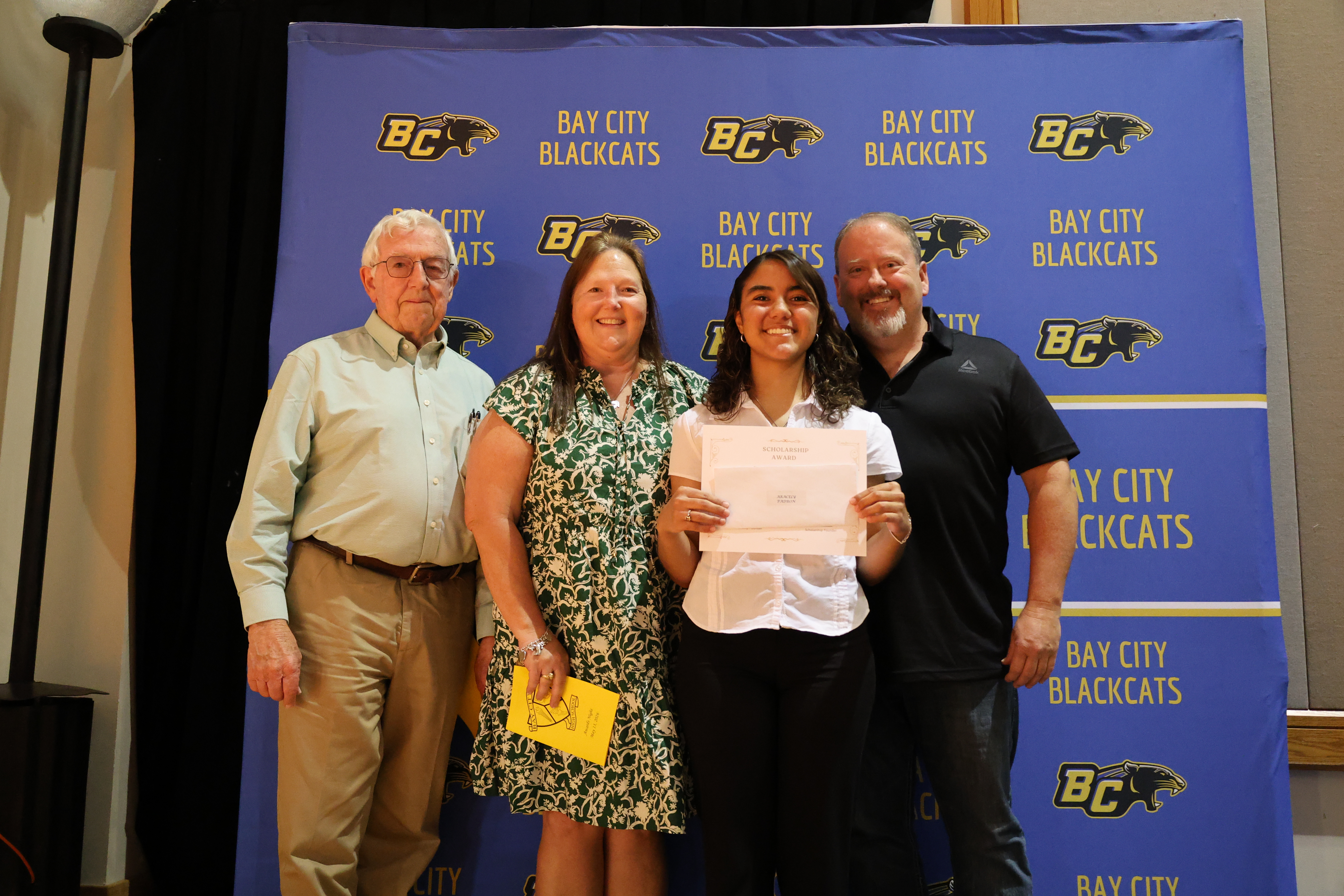 A Bailey Schmitt Memorial Fund scholarship recipient holding a certificate with Len and family in front of the Bay City Blackcats backdrop