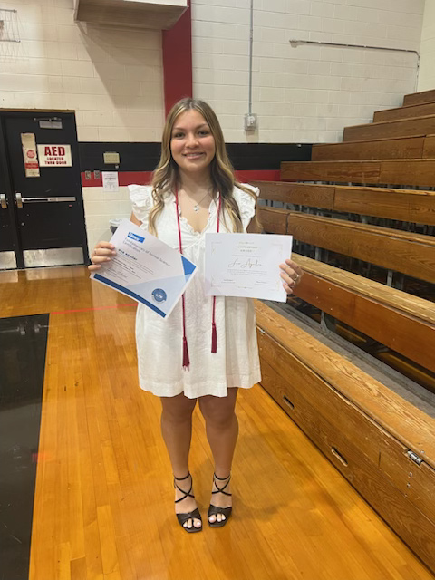 A Bailey Schmitt Memorial Fund scholarship recipient holding two scholarship certificates in a school gymnasium
