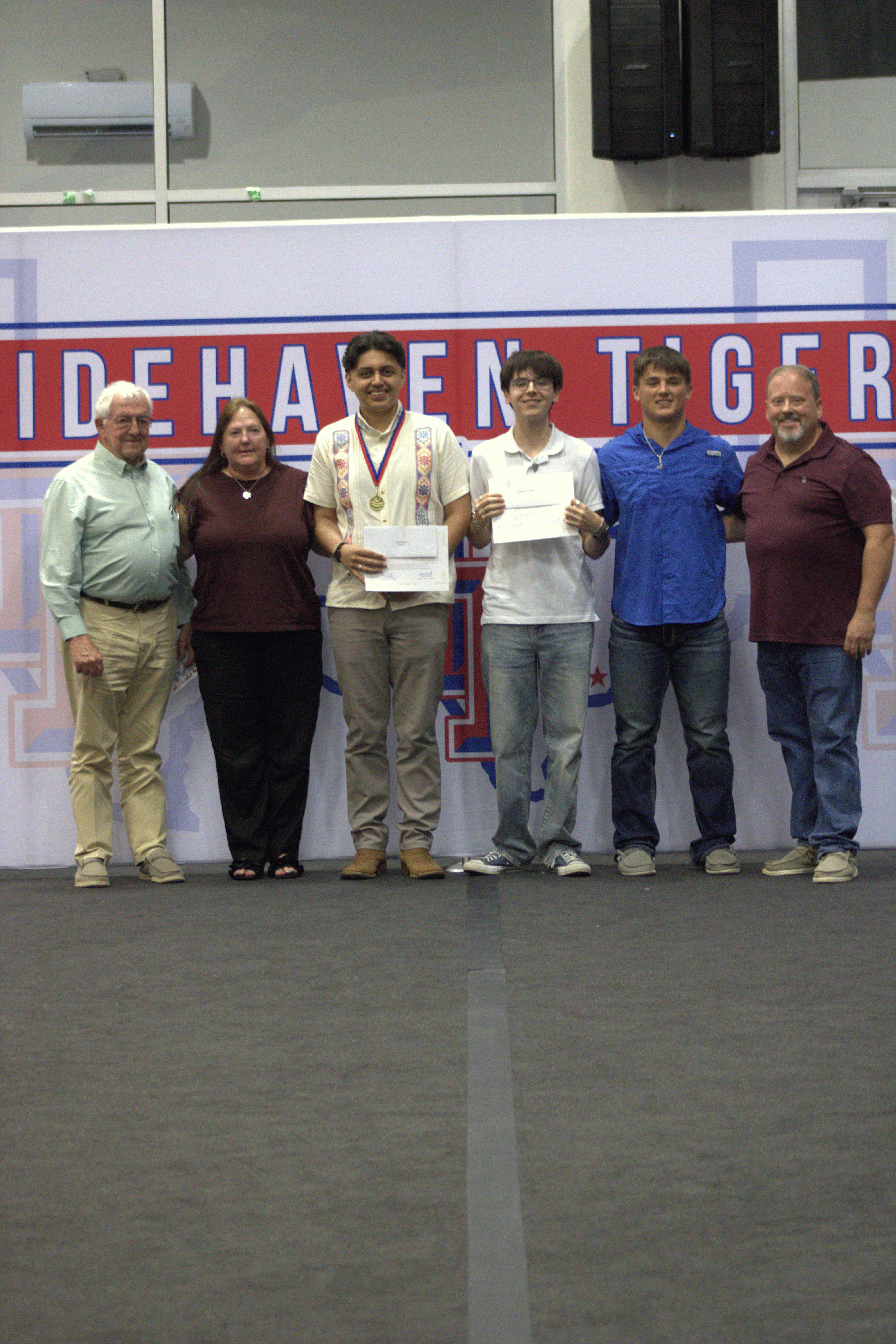 Two of the three Bailey Schmitt Memorial Fund scholarship recipients holding certificates with Len and family in front of the Tidehaven Tigers backdrop